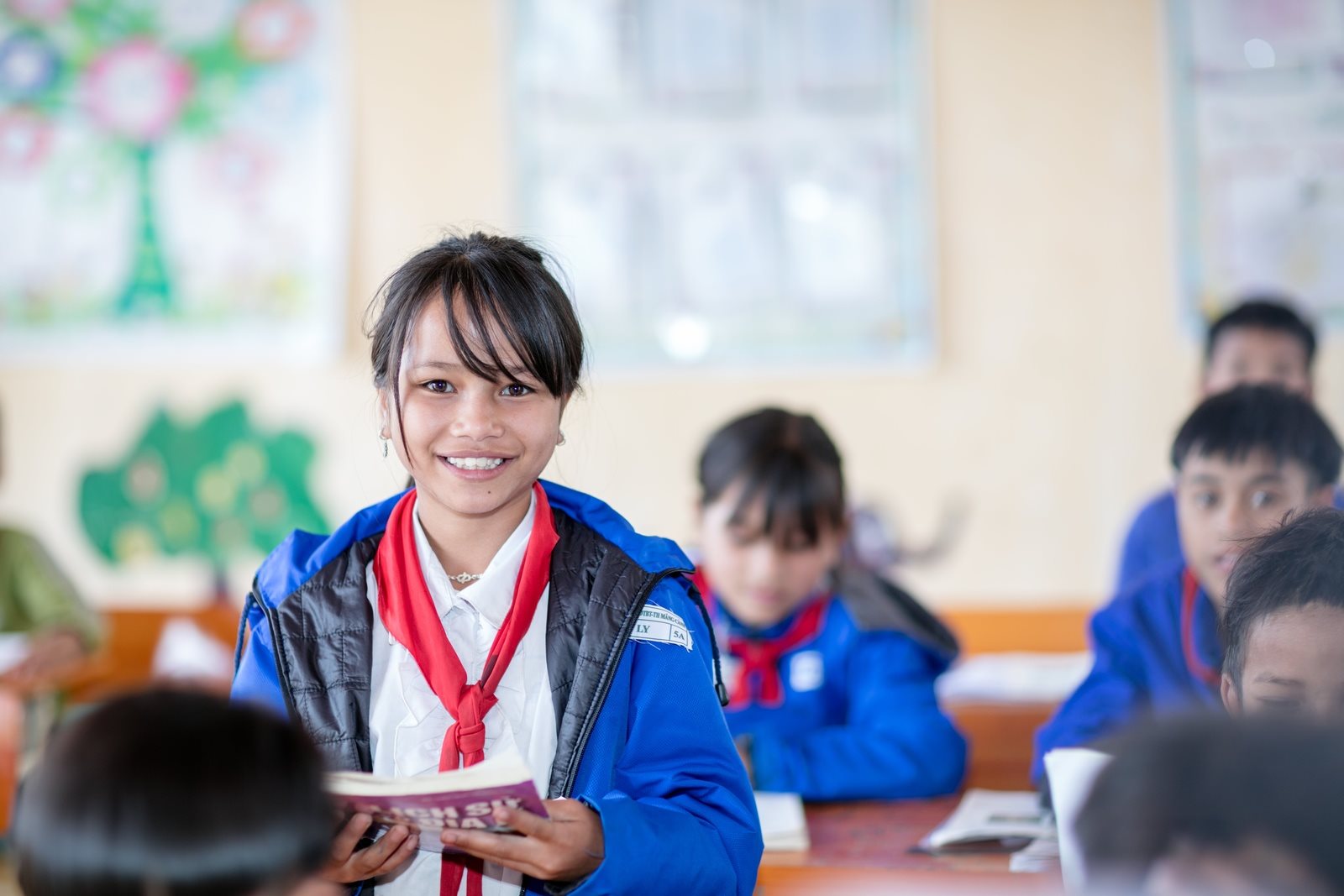 The image depicts a Latin American school classroom where both male and female students wear identical uniforms. In the foreground, a young, smiling female student stands, wearing a blue jacket and a red scarf, holding a book or notebook. Other students of similar age are seated around her. The classroom environment appears bright and colorful, adorned with artworks on the walls.