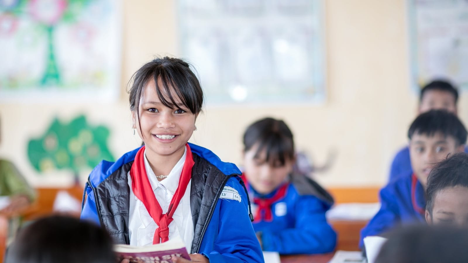 The image depicts a Latin American school classroom where both male and female students wear identical uniforms. In the foreground, a young, smiling female student stands, wearing a blue jacket and a red scarf, holding a book or notebook. Other students of similar age are seated around her. The classroom environment appears bright and colorful, adorned with artworks on the walls.