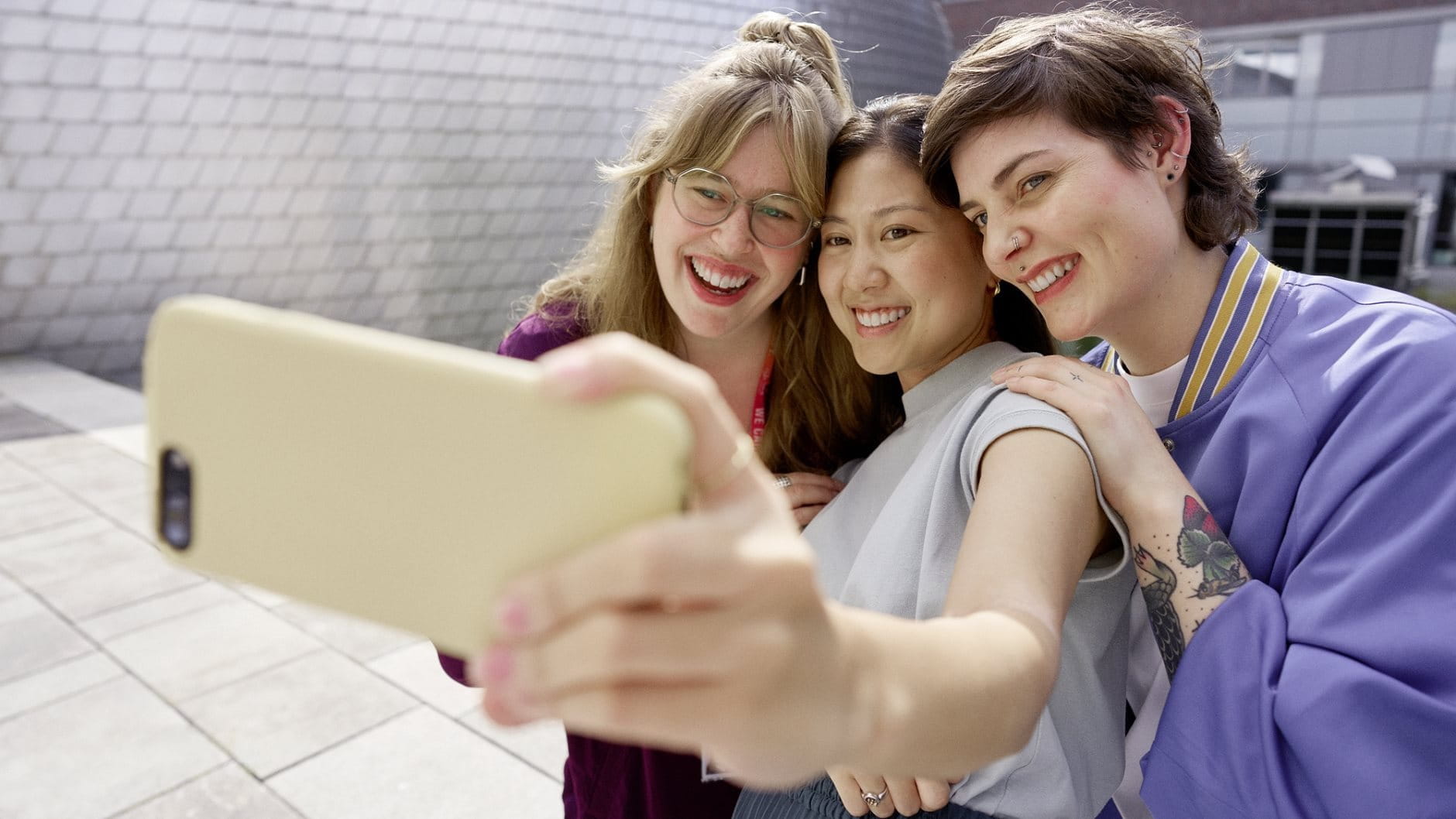 Three people smiling and taking a selfie together outdoors, holding a smartphone at arm’s length.