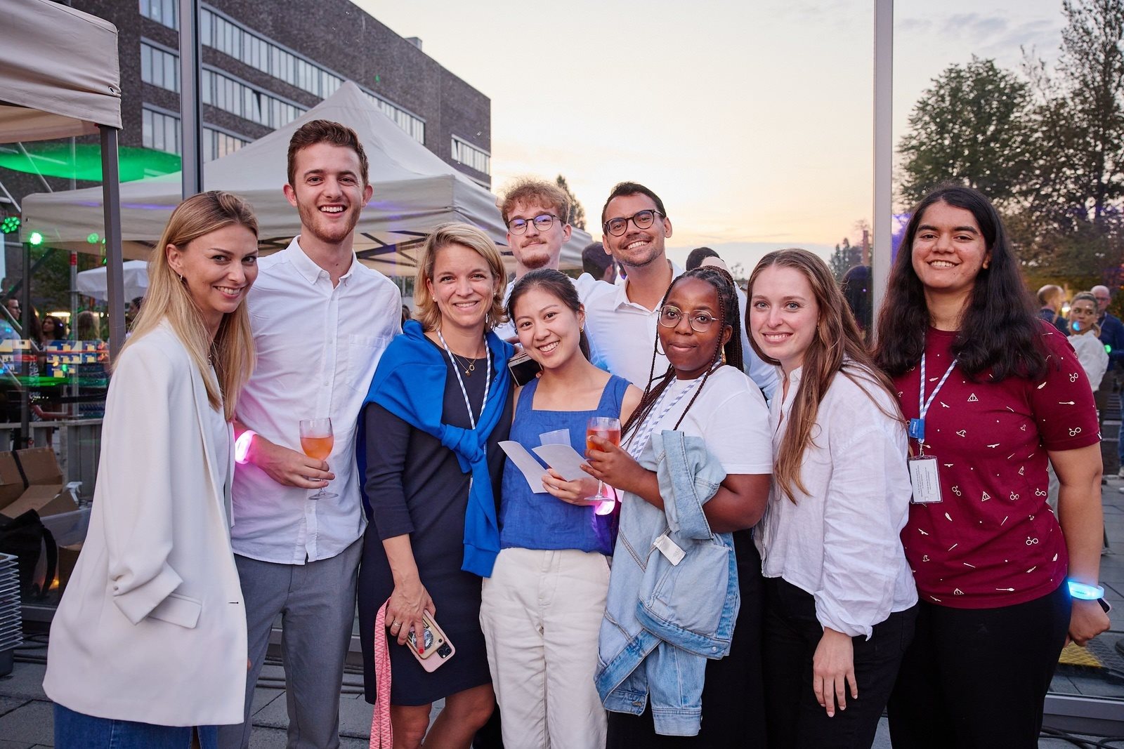 Group of young adults standing together outdoors, smiling at the camera and holding notebooks and folders on a campus setting.
