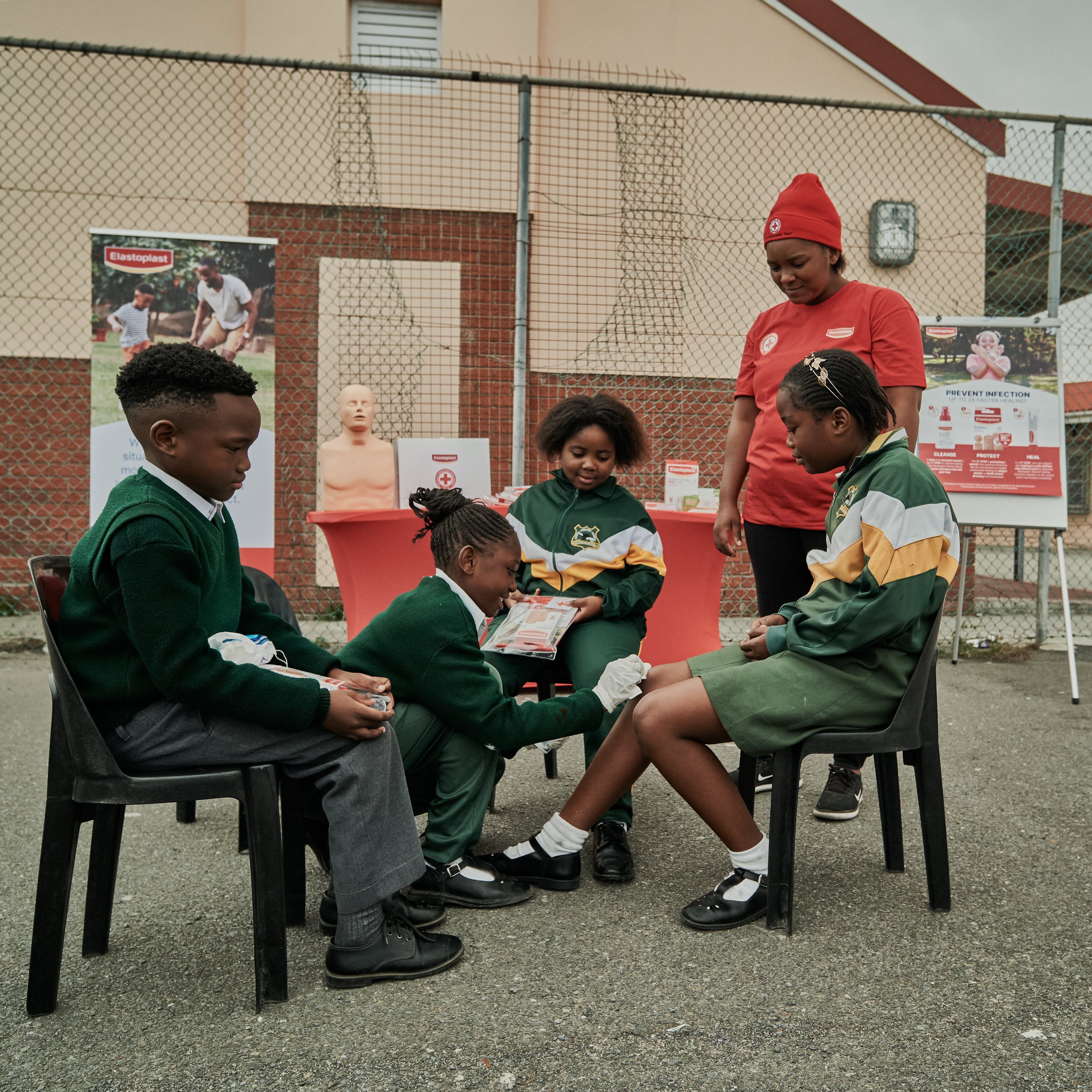 Group of schoolchildren sitting together outdoors during a first aid demonstration, with a facilitator guiding the practical learning activity.