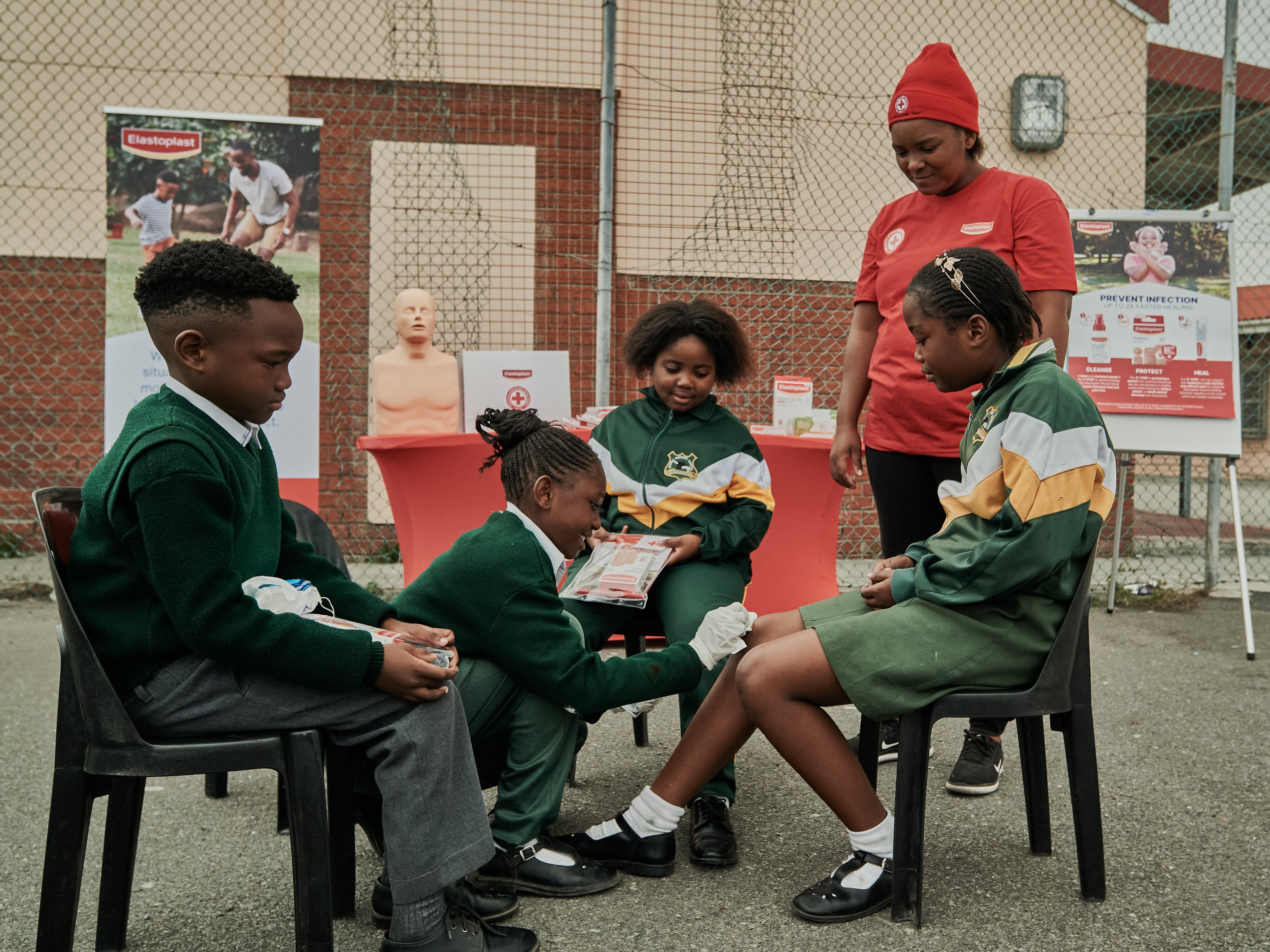 Group of schoolchildren sitting together outdoors during a first aid demonstration, with a facilitator guiding the practical learning activity.