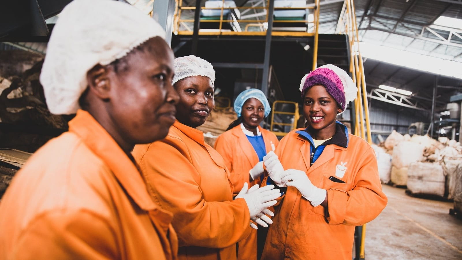 The image depicts four women with dark skin tones wearing orange work suits and bright hairnets. They are standing together in a hall or industrial setting, with large bags of collected materials visible in the background. Metal structures and scaffolding suggest a large-scale operation.