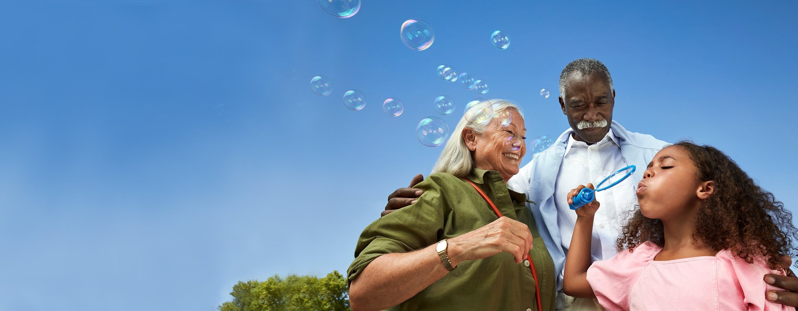 An older couple and a young child standing outdoors under a clear blue sky, smiling as the child blows soap bubbles that float around them.