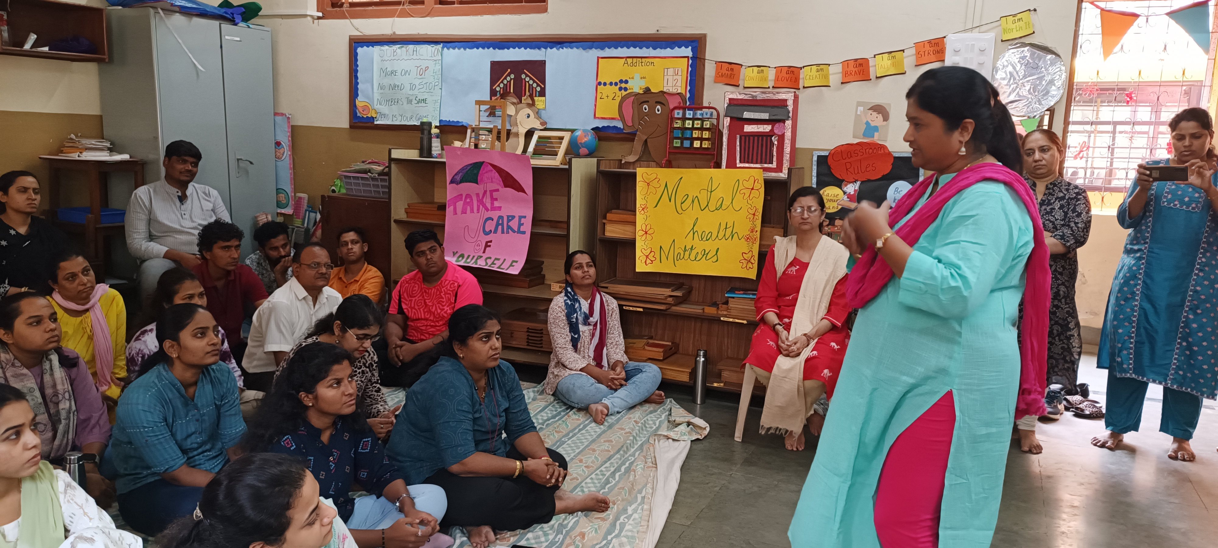 Group of girls and women sitting together in a classroom while a facilitator leads a group discussion, with posters about care, mental health, and wellbeing on the walls.