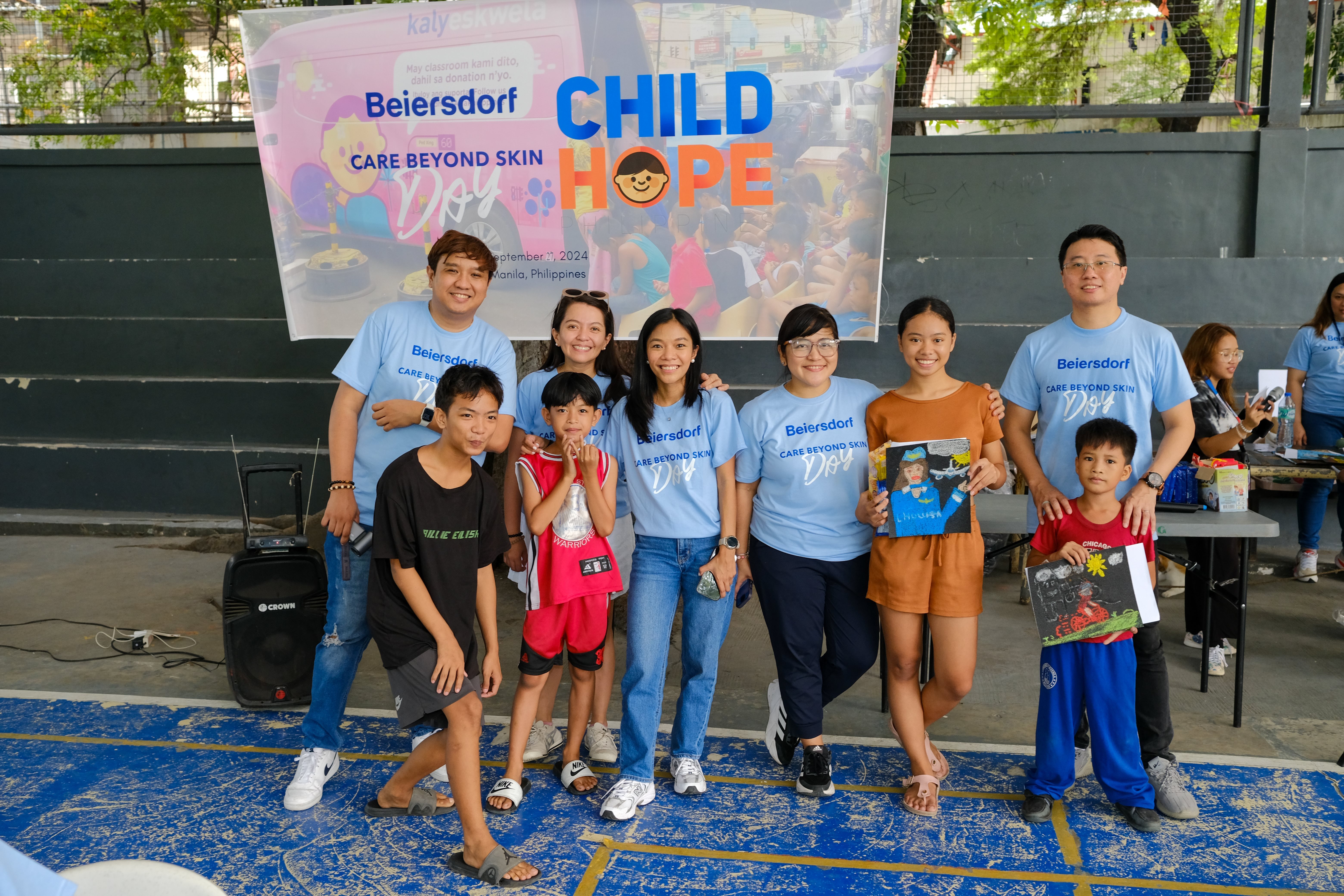 Group of Beiersdorf volunteers and children posing together at a ‘Care Beyond Skin Day – Hope’ community event, smiling and holding children’s artwork in an indoor venue.