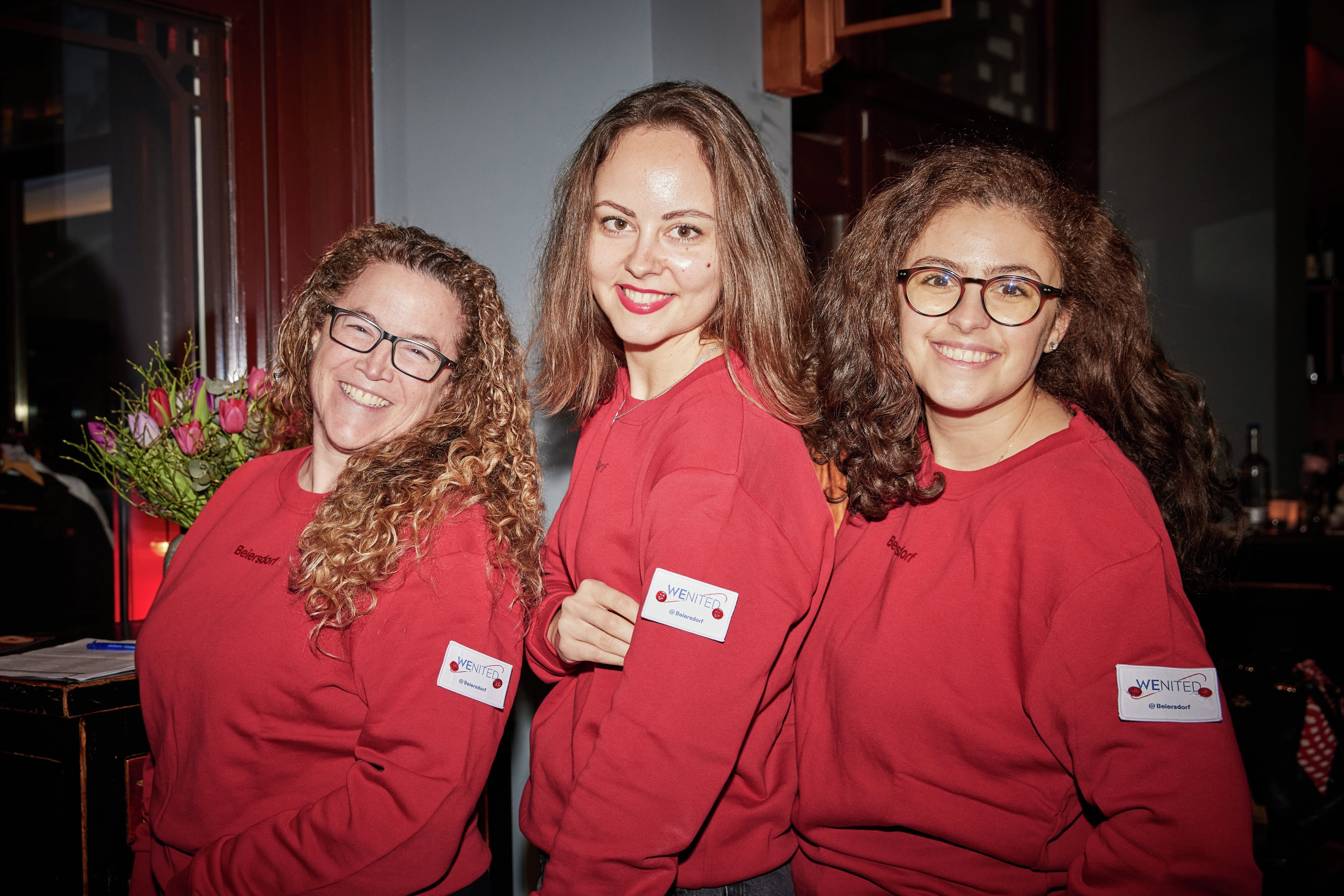 Three colleagues smiling and wearing red colored jumpers with a badge of the WEnited employee community.