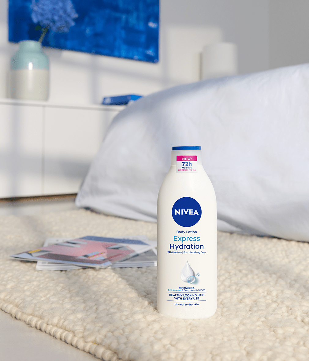 NIVEA Express Hydration body lotion bottle standing on a soft carpet in a bright bedroom setting, with a bed and magazines in the background.