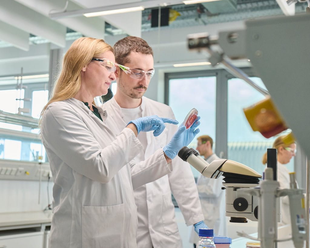 Two scientists in white lab coats and protective gloves discussing a sample in a modern laboratory, with microscopes and other researchers working in the background.