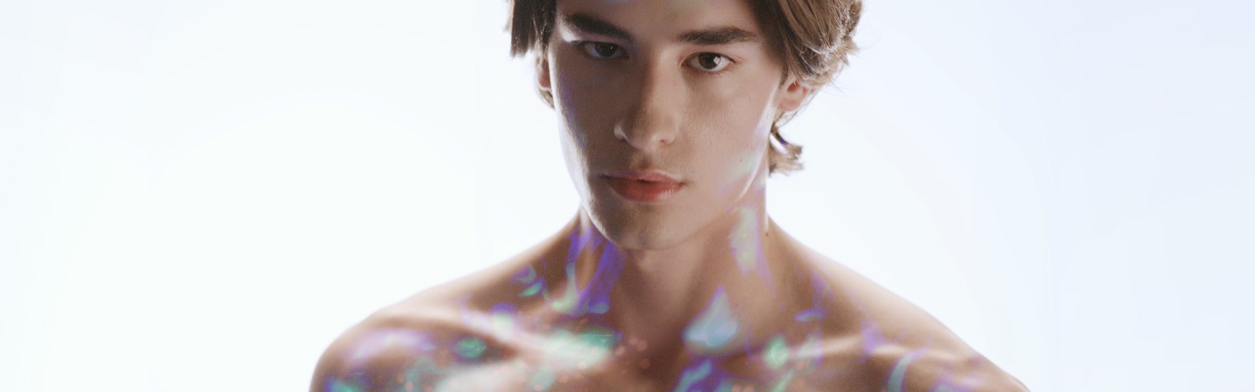 Close-up portrait of a young person against a light background, with soft, iridescent light reflections cast across their face and shoulders.