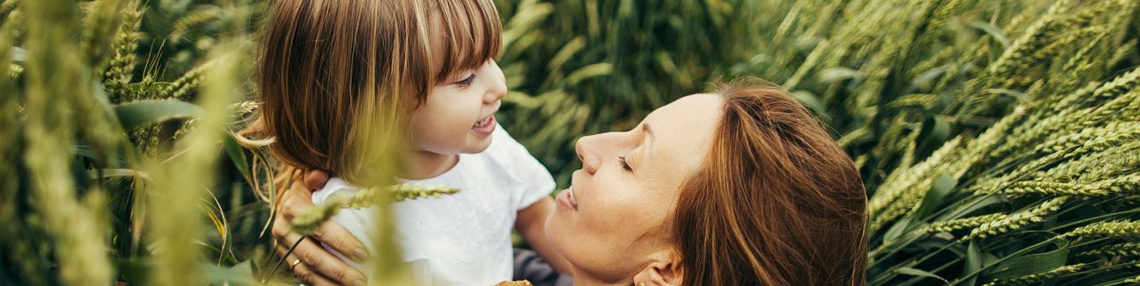 Close-up of a mother and young child smiling at each other outdoors, surrounded by greenery, conveying warmth, care, and emotional connection.