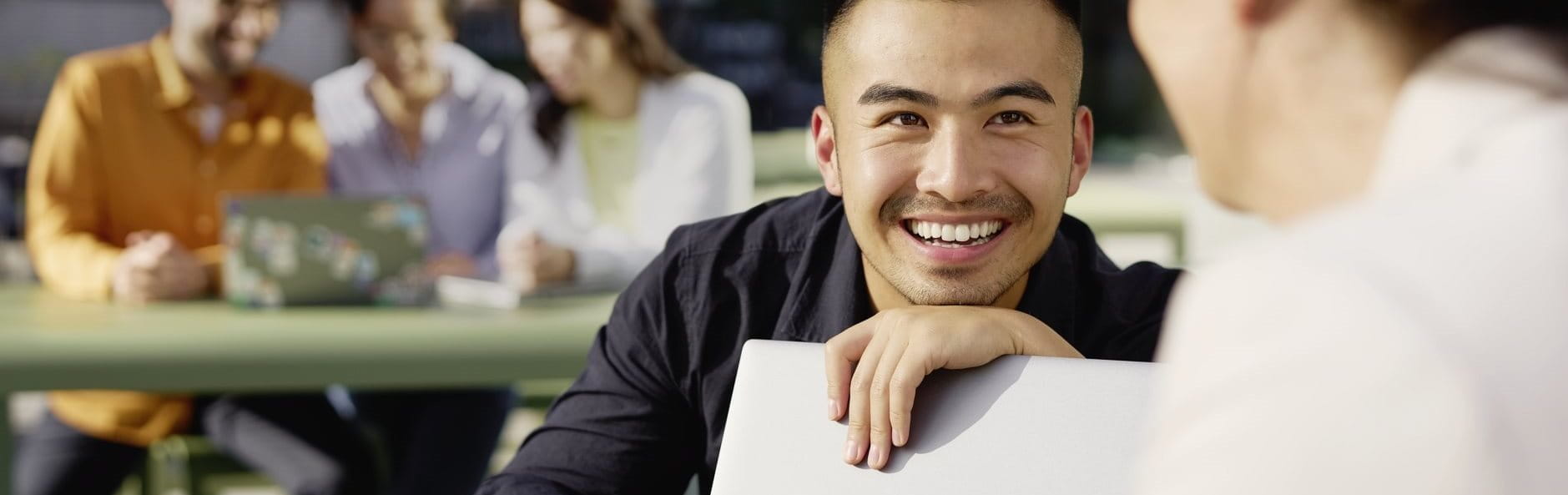 A young man smiling while talking with a colleague in a bright office or campus setting, with other people working in the background.