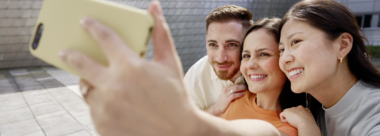Three friends taking a selfie outdoors with a smartphone, smiling together in a sunny urban setting.