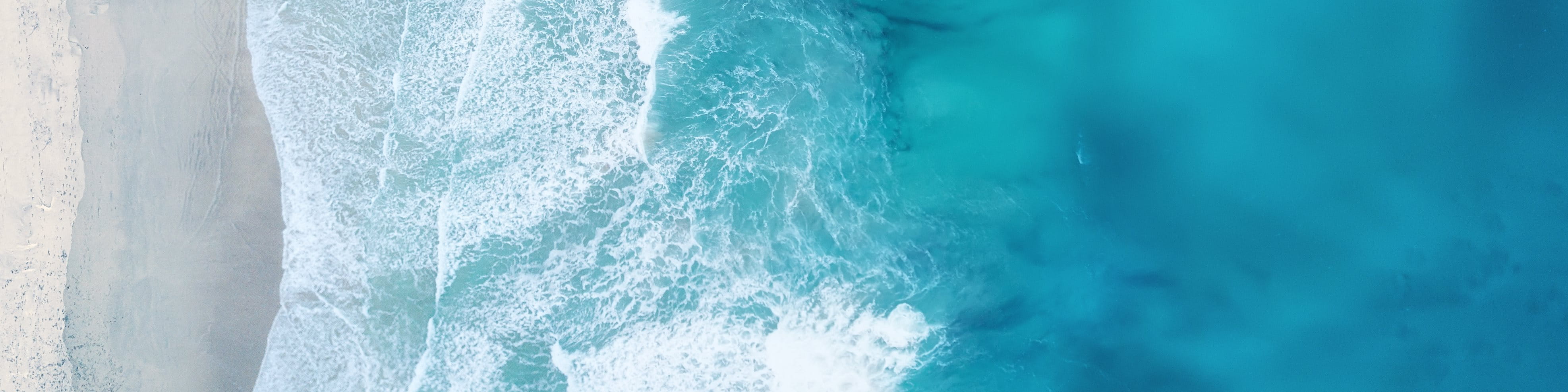 A high-angle, vertical aerial photograph looking straight down at bright turquoise ocean water meeting a light gray sandy beach. The image captures the white foam and swirling patterns of waves as they crash against the shore.